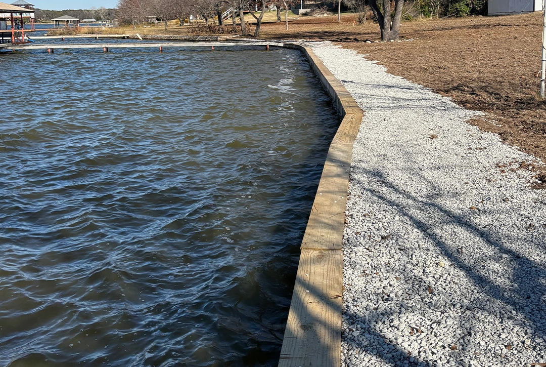 A wooden seawall or retaining wall along Lake Granbury's shoreline. The wall creates a clean barrier between the rippling water on the left and the protected shoreline on the right, which is covered with white gravel. Beyond the gravel is brown grass and scattered trees on a gently sloping property. In the distant background, you can see other lakefront properties and docks. The image showcases shoreline erosion protection on a winter or early spring day.
