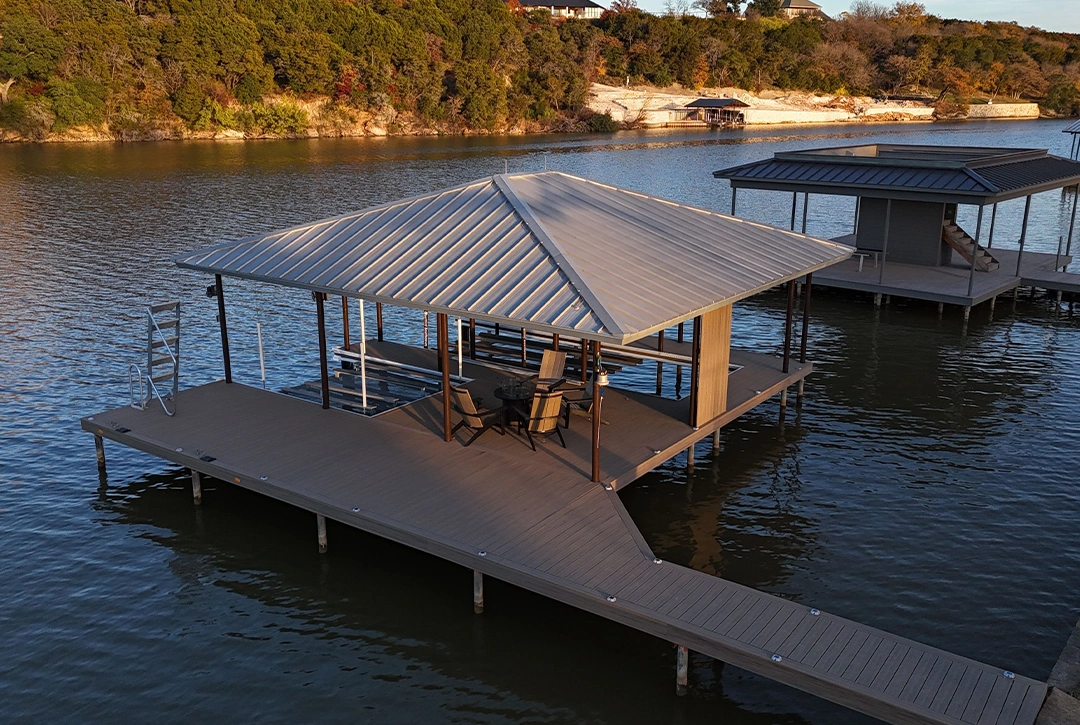 A custom boat dock extends into Lake Granbury with a modern design featuring a covered slip area with a metal hip roof. The dock has composite decking in a warm brown tone and includes a seating area with wooden Adirondack chairs. A long walkway connects the main platform to the shore. In the background, another boat dock is visible across the calm water, and the shoreline is lined with trees and natural vegetation. The scene is captured during golden hour, with warm sunlight highlighting the metal roof and creating a peaceful lakeside atmosphere. Hills with residential properties can be seen in the distance.