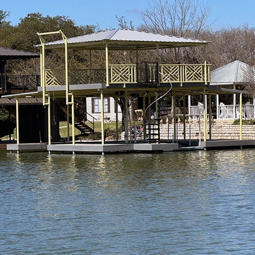 A charming two-story yellow dock on Lake Granbury featuring intricate Chippendale handrails and a spiral staircase. The cottage-inspired waterfront structure showcases custom-fabricated geometric fretwork panels, creating an elegant and unique retreat in the Holiday Estates neighborhood.