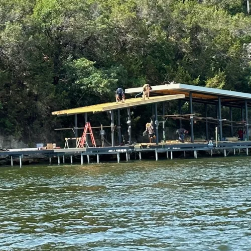 Construction workers repairing a boat dock on Lake Granbury. Several people are visible on the metal frame structure installing new decking on both the main platform and upper level. The dock extends from a tree-lined shore into calm water, with construction equipment including ladders visible on the platform.
