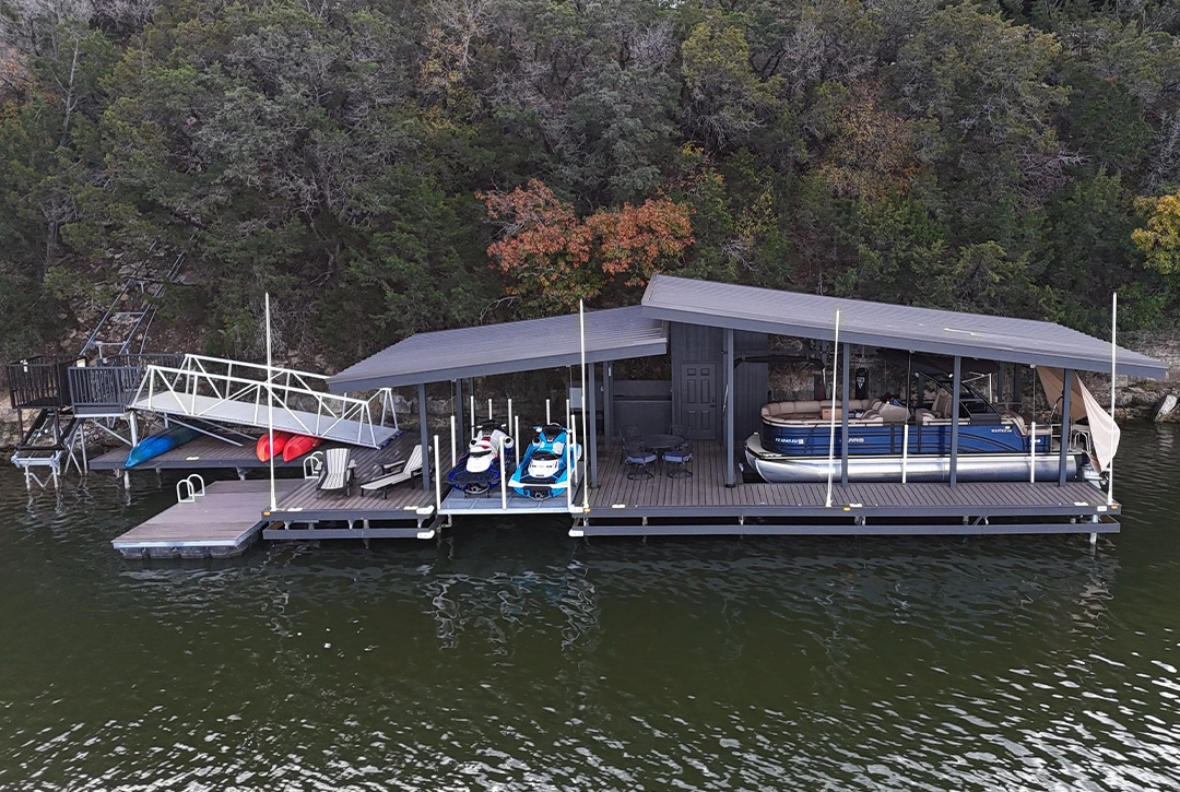An aerial view of a covered boat dock on Lake Granbury with a gray metal roof and composite decking. The dock features multiple slips housing various watercraft including a blue pontoon boat, jet skis, and smaller vessels. A storage enclosure with a door is built into one side of the structure. The dock includes an access gangway with white railings connecting to the shore. Dense trees in fall colors surround the shoreline, creating a natural backdrop for the waterfront structure.