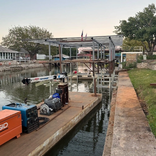 A partially constructed boat dock in a canal, featuring a steel frame for the roof and dock supports. Construction equipment, including a generator and welding tools, is visible on the floating work platform, indicating an ongoing installation process.