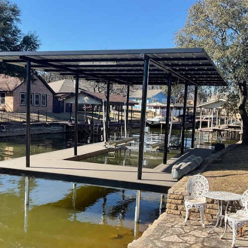A modern black steel-framed boat dock with a sleek metal roof and wooden decking, positioned in a peaceful canal. A white outdoor bistro set sits nearby on the shore, enhancing the lakeside relaxation atmosphere.