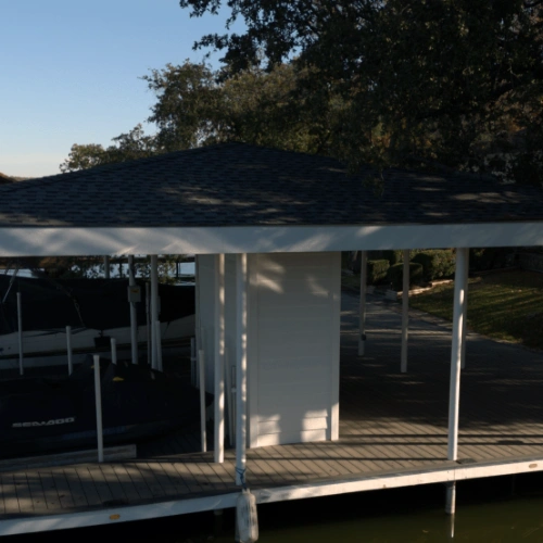 A completed Polynesian boat dock with a covered structure featuring a shingled roof and a dedicated enclosed storage room. The dock has ample space for multiple watercraft, including a Sea-Doo personal watercraft and a boat lift, providing both protection