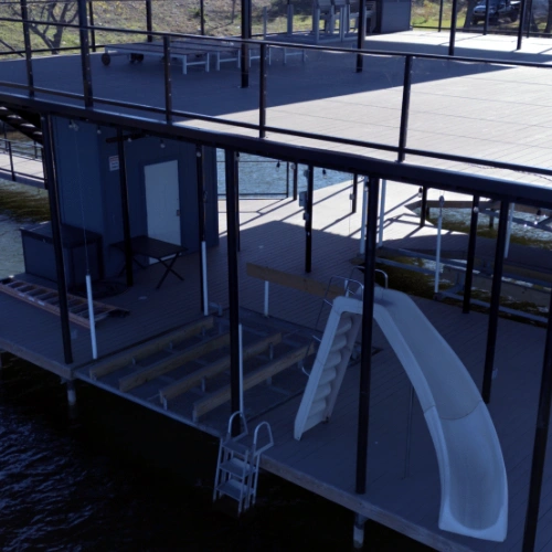 A luxurious two-story Polynesian boat dock featuring an upper-level entertainment deck with a black metal railing. The lower level includes covered boat slips, an enclosed storage room, and a built-in waterslide leading into the lake. A ladder provides ea
