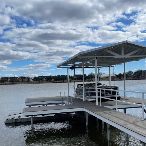 A partially constructed boat dock overlooking a scenic lake. The framework of the dock is in place, with a gabled metal roof supported by steel posts. The spacious platform extends over the water, offering a glimpse of its future entertainment potential.